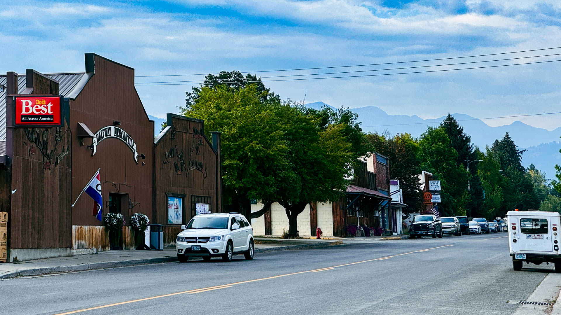 Halfway OR 2 A small-town street scene in Halfway, OR, with cars parked along the road, a white SUV driving, rustic buildings, a Best All home brands sign, trees, and mountains visible in the background under a blue sky with clouds.