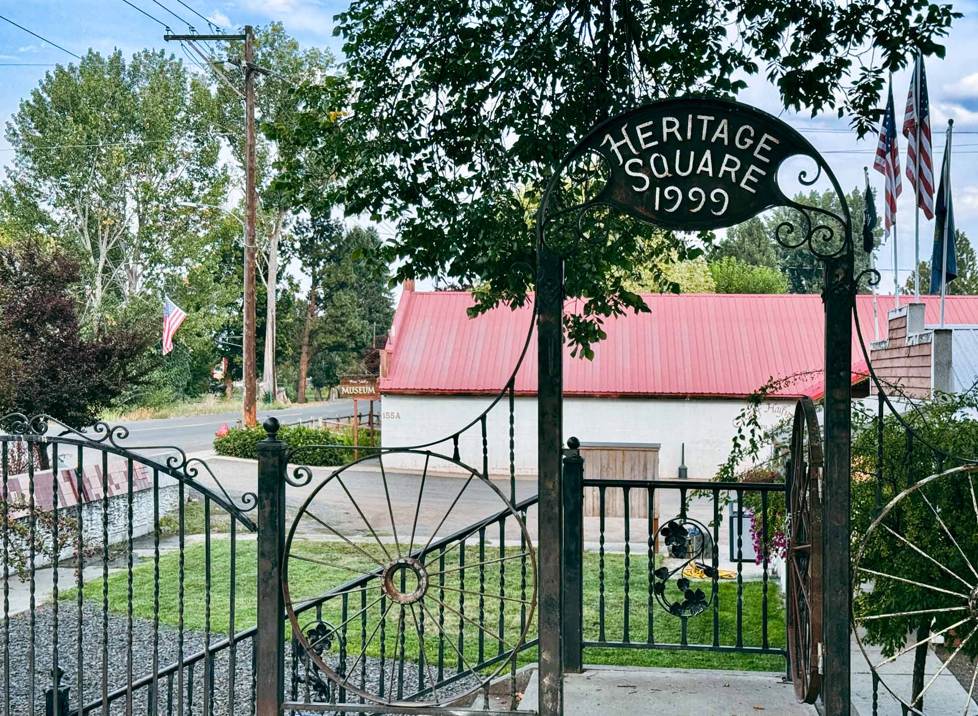 Halfway OR 4 An ornate black metal gate reading Heritage Square 1999 stands at the entrance, beckoning visitors to the garden area in Halfway OR, with a red-roofed building and flags visible beneath a partly cloudy sky.
