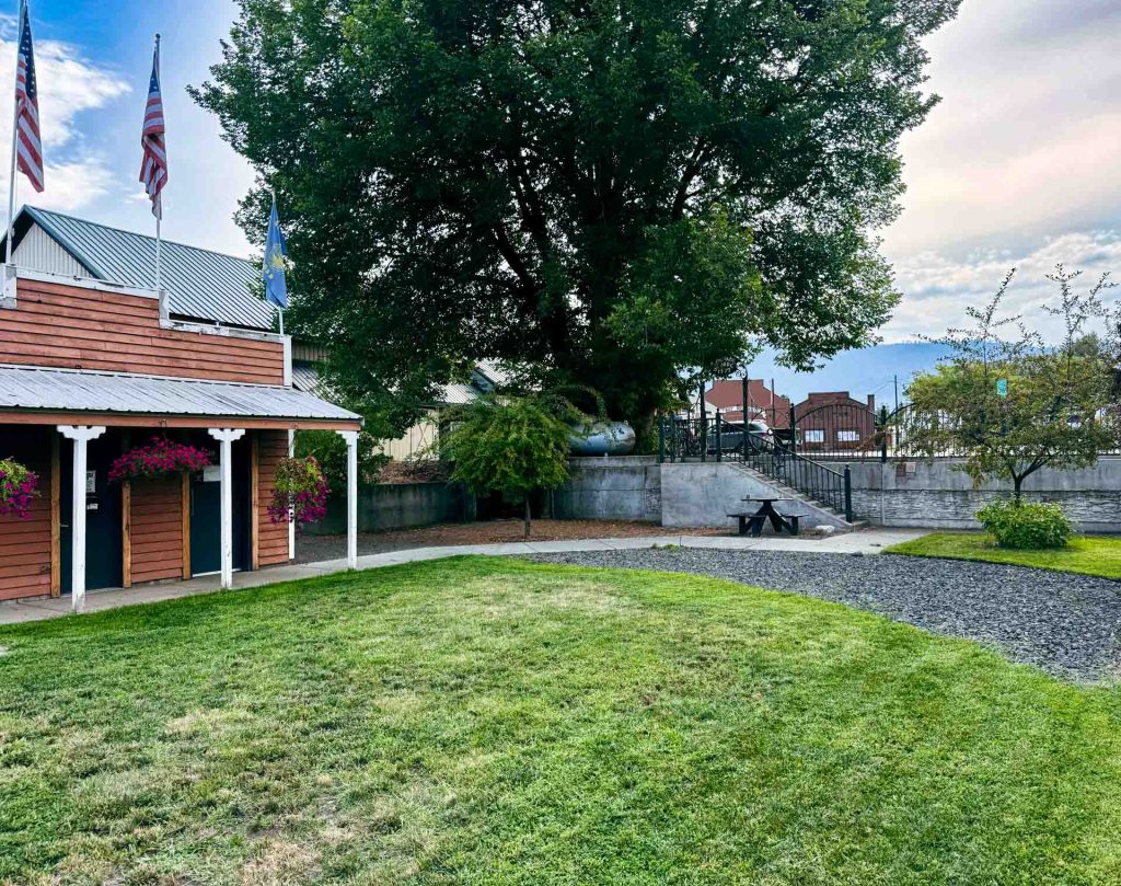 Halfway OR 11 A grassy yard in Halfway OR features a wooden building on the left, hanging flower baskets, flags on poles, a large tree at the center, a picnic table, and buildings in the background under a partly cloudy sky.