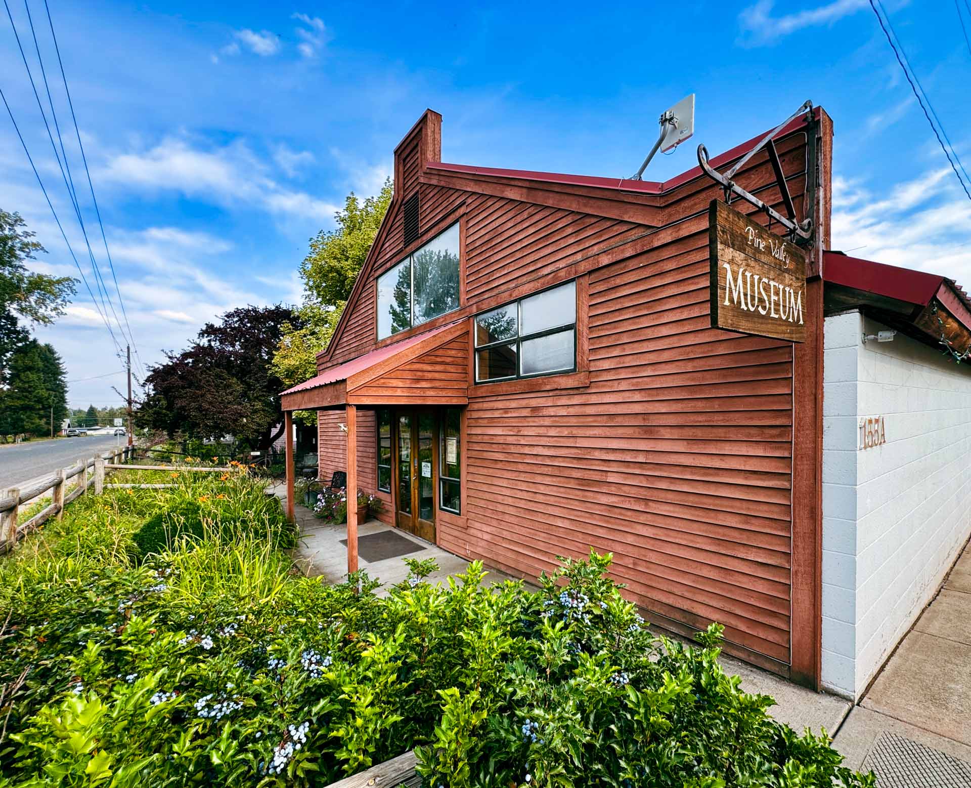 Halfway OR 3 A rustic wooden building in Halfway, OR, displays a sign reading Museum near the entrance. Lush greenery and a sidewalk border the front, with a road and trees visible on the left under a partly cloudy sky.