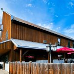 Halfway OR 13 A rustic wooden restaurant in Halfway, OR with a sign reading The Main Place, red patio umbrellas, outdoor seating, a wooden fence, and a bright blue sky with clouds above.