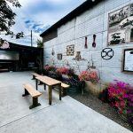 Halfway OR 17 Outdoor seating area with wooden benches and tables beside a white brick wall decorated with western-themed art, signs, and flowers in hanging baskets; a food truck is visible in the background.