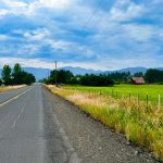 Halfway OR 25 A rural road in Halfway, OR runs alongside green fields with a red barn, fencing, and distant forested mountains under a partly cloudy sky. Power lines and trees border the scene.