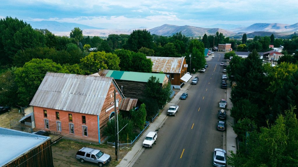 Halfway OR 26 Aerial view of Halfway OR shows a small town street lined with trees, parked cars, and rustic buildings, set against a backdrop of mountains and cloudy skies.