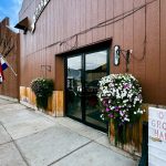 Halfway OR 16 A brown building in Halfway, OR, with a glass door, flower pots, and a sign that reads “OPEN GROCERIES HARDWARE.” An American flag hangs near the door while a “Best” sign is visible down the empty sidewalk.