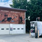 Halfway OR 18 A building in Halfway OR with a mural of wildlife and mountains, labeled Halfway Mercantile, stands beside a fuel tank and gas pump. Trees and a cloudy sky form the scenic background.