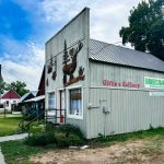 Halfway OR 20 A rustic gray wooden building in Halfway, OR, adorned with animal carvings and signs reading “Gifts & Gallery” and “Farmer’s Market,” sits along a small-town street with a church and trees under a partly cloudy sky.